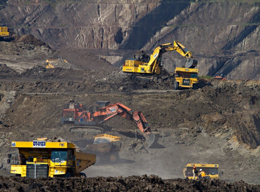 A wide-angle, high-perspective photograph of a vast open-pit mining operation. Multiple pieces of heavy industrial machinery are active across tiered dirt terraces, including large yellow haul trucks and hydraulic excavators in yellow and orange. The scene captures the scale of resource extraction, with steep, excavated rock walls in the background and a dusty, industrial atmosphere.